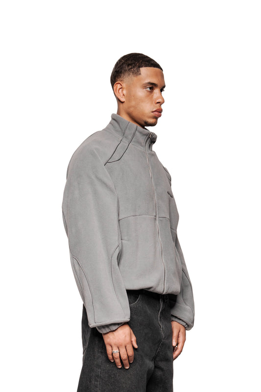 Junger Mann im Profil mit Fleece Jacket Night Owl, graues Zip-up mit OLA KALA-Logo, Teilungsnähten, Eingriffstaschen, und elastischen Bündchen, präsentiert minimalistischen Stil.