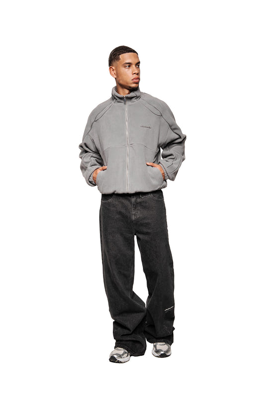 Young man in casual street style, wearing the Night Owl fleece jacket with OLA KALA embroidery, slit pockets and elasticated cuffs, combined with wide jeans and sneakers.