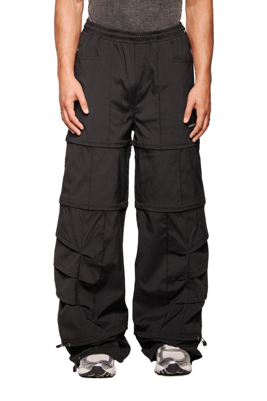 Person trägt Gills Zip-Off Pants Black, mit abnehmbaren Beinteilen, Kiemen-Lufteinlässen, mehreren Taschen und gesticktem OLAKALA-Logo, kombiniert mit grauen Sneakers.