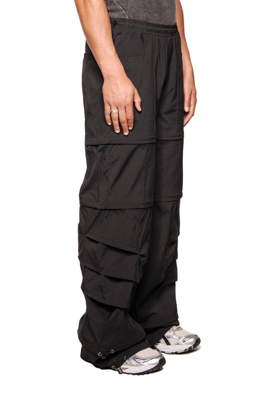 Modell trägt Gills Zip-Off Pants Black mit mehreren Taschen, abnehmbaren Beinteilen und Kordelsäumen, kombiniert mit silbernen Sneakers, Teil der OLAKALA-Kollektion.