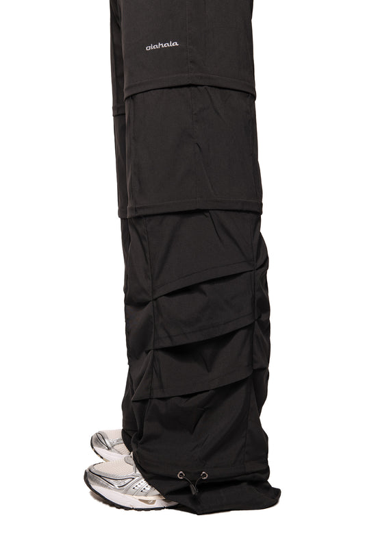 Close-up of Gills Zip-Off Pants Black with detachable leg sections, embroidered OLAKALA logo on the calf and silver and white sneakers, elastic cord at the hem.