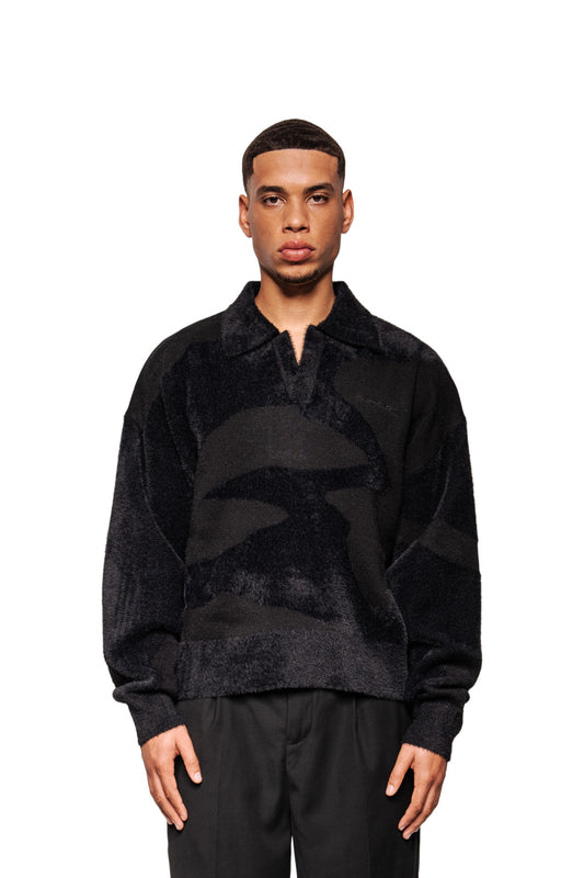 Männermodell trägt den Glitched Fuzzy Polo Black, ein unisex Pullover mit Polo-Kragen, abstraktem Muster, gesticktem Logo und Ripp-Bündchen, passend zu schwarzen Hosen.