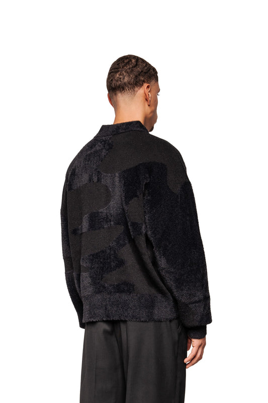 Person wears Glitched Fuzzy Polo Black, a unisex, fluffy, black polo with polo collar, no buttons and embroidered Synergy logo, visible from the back.