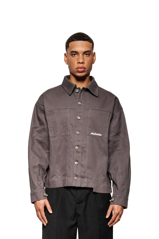 Junger Mann in grauer Heavy Anatomy Cotton Overshirt von OLAKALA mit Knopfleiste, Brusttaschen und gesticktem Logo, über schwarzen Hosen getragen.