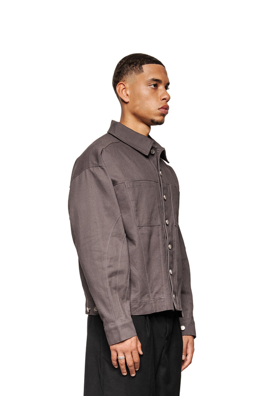 Männliches Modell im Profil trägt Heavy Anatomy Cotton Overshirt Gray mit Knopfleiste, gesticktem OLAKALA-Logo und lockerer Passform, kombiniert mit schwarzen Hosen.