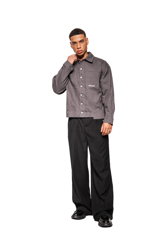 Men's model wears the Heavy Anatomy Cotton Overshirt Gray with button placket, breast pockets, embroidered OLAKALA logo and dividing seams, combined with black, wide-cut pants.