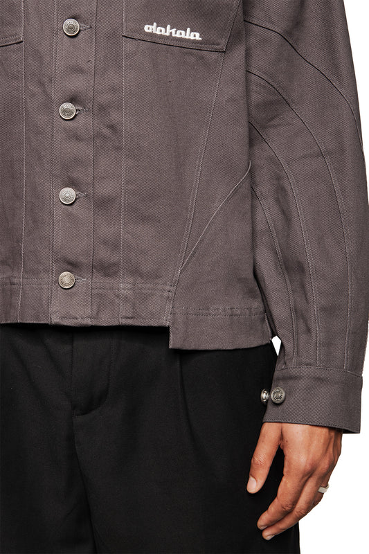 Person wears Heavy Anatomy Cotton Overshirt Gray with silver press studs, embroidered OLAKALA logo on the breast pocket and dividing seams, combined with black pants.