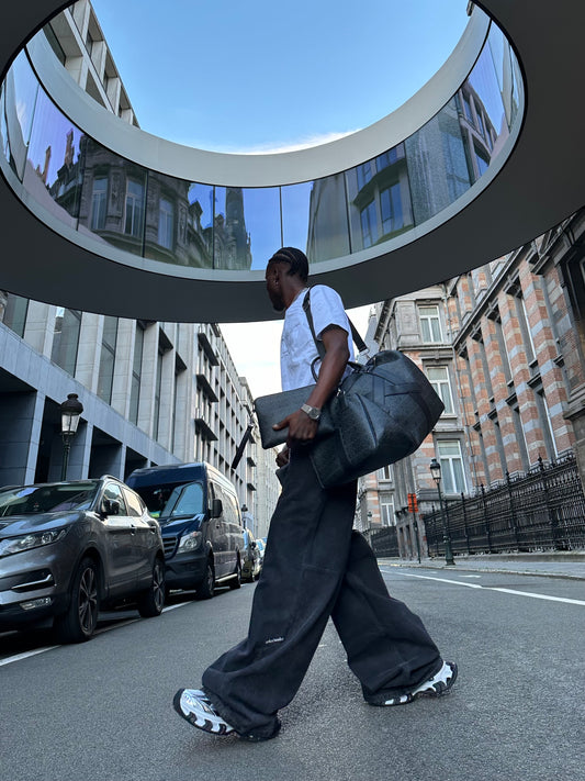 A man in a white T-shirt and loose black pants is walking down an urban street; he is carrying a large dark duffel bag and a flat laptop sleeve, beneath a round glass canopy.