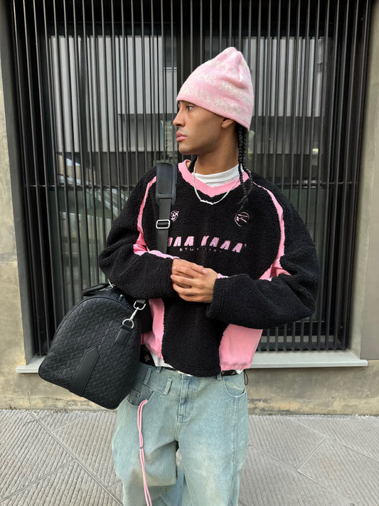 A person wearing a pink beanie and a black fleece sweater with pink accents, light-colored baggy jeans with a pink drawstring, and a black shoulder bag – against an urban backdrop.