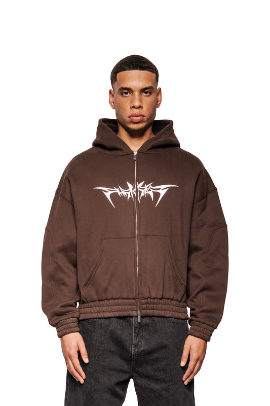 Männliches Model trägt Isolation Zip Hoodie Brown mit weißem Tribal-Print, Kängurutaschen und elastischen Bündchen. Entspannte Haltung, zeigt Oversized-Unisex-Design.