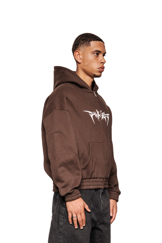 Männliches Modell trägt Isolation Zip Hoodie Brown mit Kängurutasche, Foam-Print-Logo, elastischen Bündchen und Oversized-Passform, kombiniert mit dunklen Jeans.