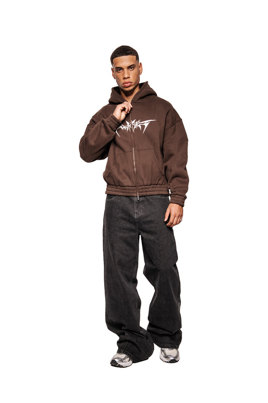 Junger Mann im Isolation Zip Hoodie Brown mit Foam-Print, elastischen Bündchen, Fronttaschen, und 2-Wege-Reißverschluss, kombiniert mit schwarzen Jeans und Sneakers.