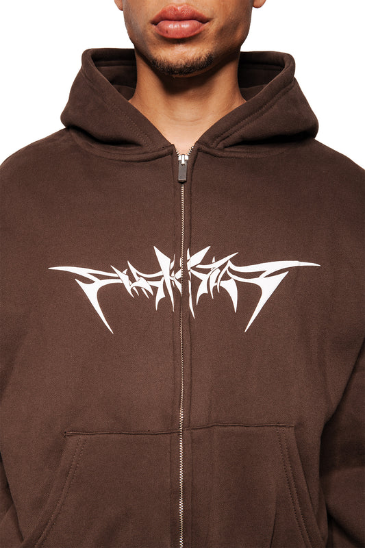 Person trägt braunen Isolation Zip Hoodie mit stilisiertem Foam-Print auf der Brust, Kapuze zurückgeschlagen, sichtbare Kängurutasche, elastische Bündchen, Oversized-Passform.