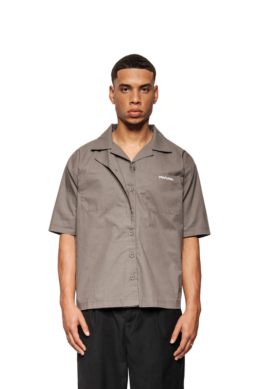 Modell trägt Layered Short Sleeve Shirt Gray mit kubanischem Kragen, Knopfleiste, gesticktem OLAKALA-Logo auf der Brusttasche und lockerer Passform.