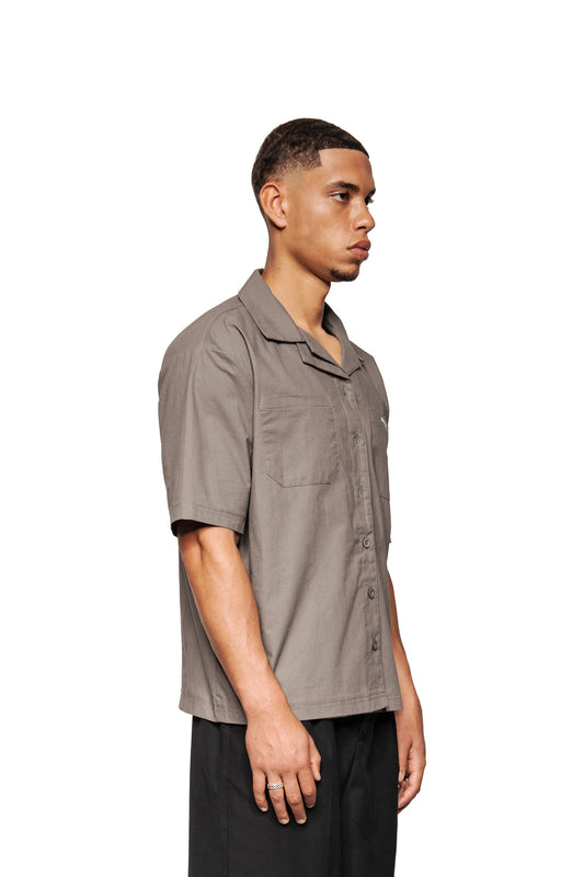 Junger Mann im Profil trägt ein graues Layered Short Sleeve Shirt mit kubanischem Kragen und OLAKALA-Logo auf der Brusttasche.