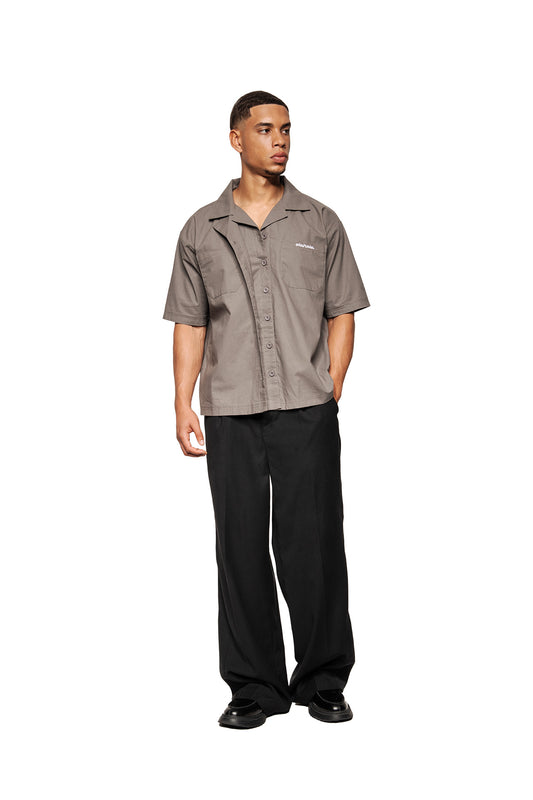 Male model wears the Layered Short Sleeve Shirt Gray with Cuban collar and embroidered OLAKALA logo, combined with black, wide-cut pants and chunky leather shoes.