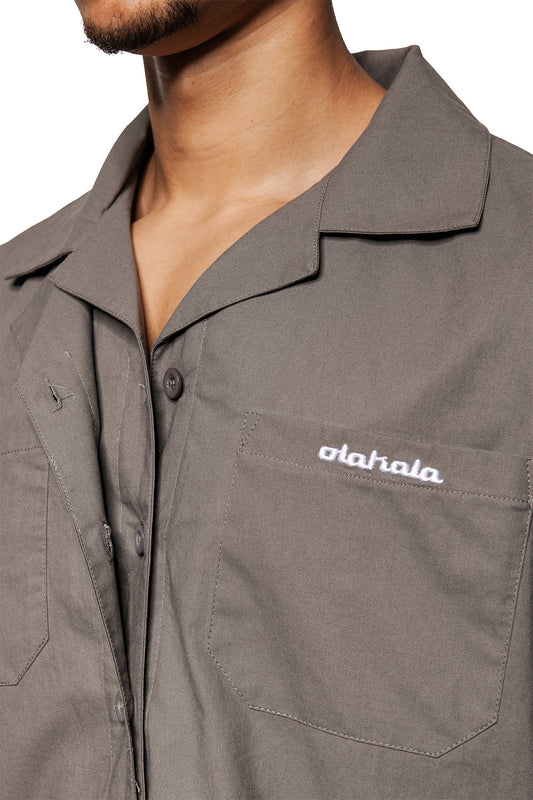 Person wears a Layered Short Sleeve Shirt Gray with Cuban collar, button placket, chest pocket and embroidered OLAKALA logo. Loose, unisex cut.