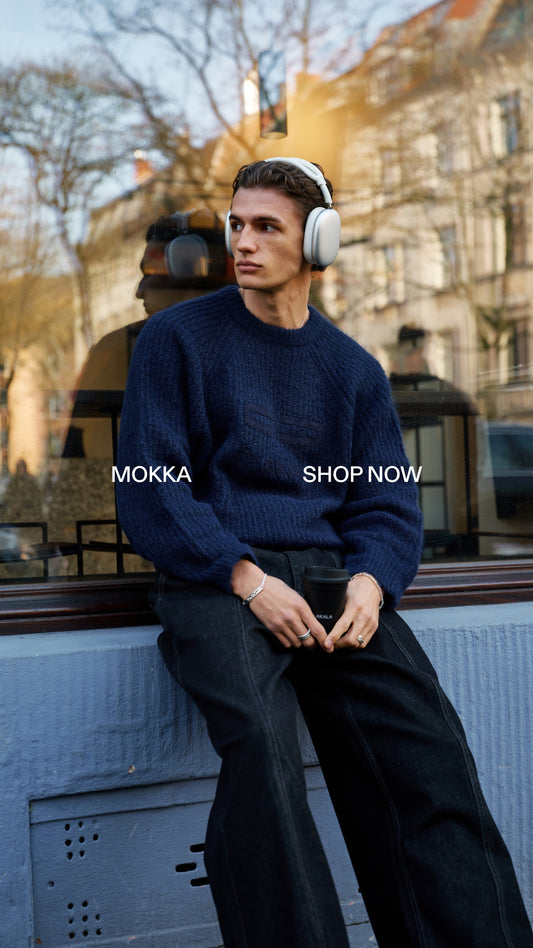 Junger Mann in dunkelblauem Grobstrickpullover und schwarzen Jeans sitzt draußen auf einer blauen Fensterbank, trägt weiße Kopfhörer und hält einen To-go-Kaffee-Becher; Reflektionen im Glas hinter ihm.