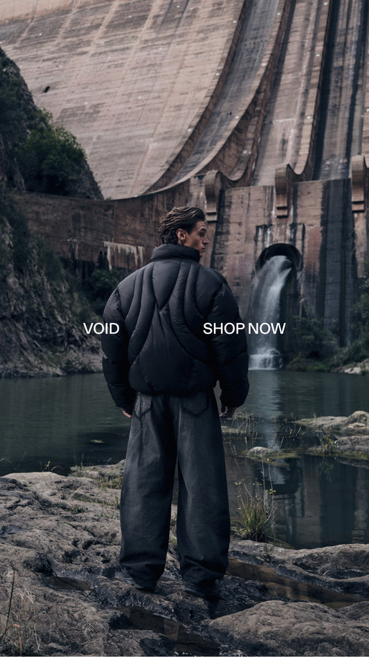 Person von hinten in schwarzer Daunenjacke und dunkler Hose steht am Flussufer vor einer Staumauer mit Wasserfall; Werbebotschaft „VOID“ und „SHOP NOW“ sichtbar.