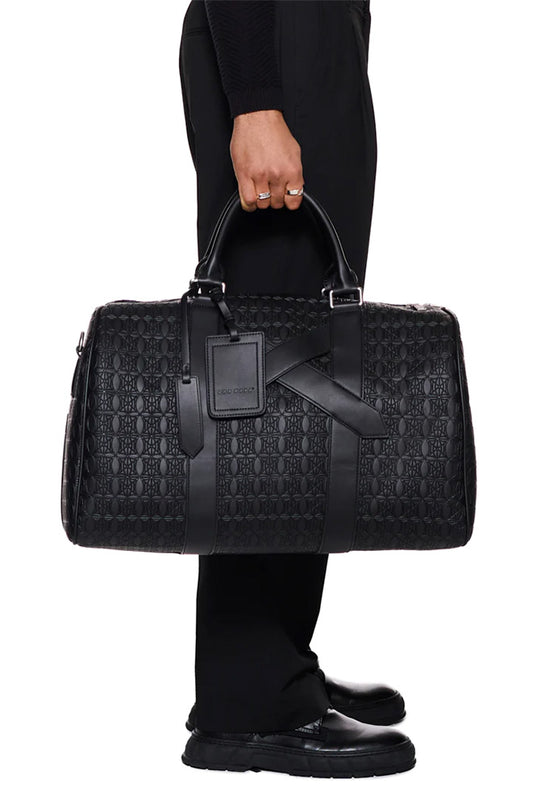 Person hält CC Debossed Weekender Bag Black mit OLAKALA Monogramm, abnehmbaren Schulterriemen und Metallreißverschluss.