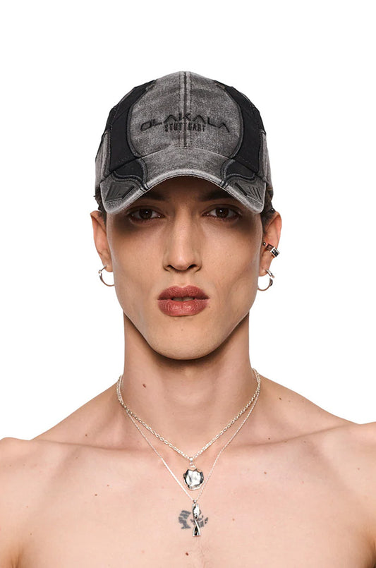 Synergy Motocross Cap mit OLA KALA-Stickerei, verstellbarem Riemen, Gummi-Patches auf dem Schirm, getragen von einem Mann mit Ohrringen und Halskette.