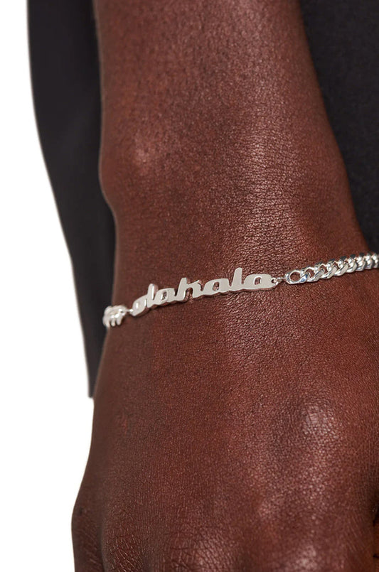 Person trägt das CC Logo Armband Silber beschichtet mit OLA KALA Anhänger und Cuban Link Kette, 0,5 cm Breite, 19 cm Länge.