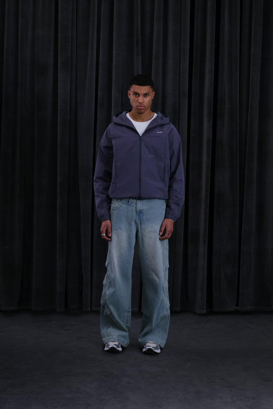 Vollständige Körperansicht eines Mannes in dunkellila Windbreaker mit Kapuze, weißem T‑Shirt, locker sitzender Jeans und Sneaker vor schwarzem Vorhang.