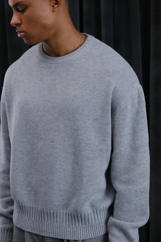 Hellgrauer Rundhals-Pullover aus weichem Strick, gerippter Saum und Bündchen, dezentes Logo auf der Brust.