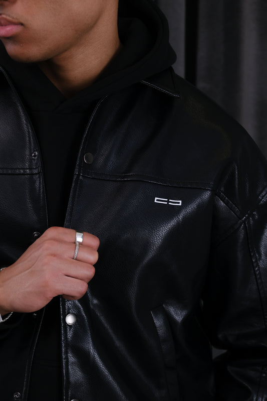 Nahaufnahme einer Person in schwarzer Lederjacke mit kleinem weißen Logo links auf der Brust, über einem schwarzen Hoodie; eine Hand mit Silberring greift an den Knopf.