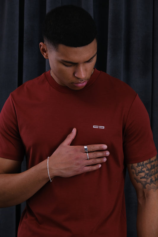 Mann in rotem T‑Shirt mit kleinem weißen Logo auf der Brust, Hand auf der Brust, silberner Ring und Armband, sichtbares Tattoo am Unterarm, dunkler Hintergrund.