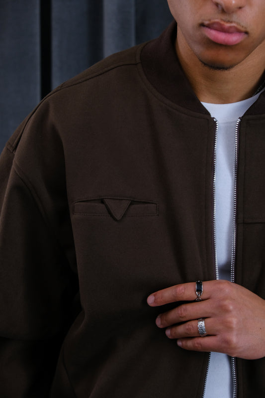 Nahaufnahme einer dunkelbraunen Reißverschluss-Jacke mit Fronttasche und Dreiecksdetail, über einem weißen Shirt getragen.