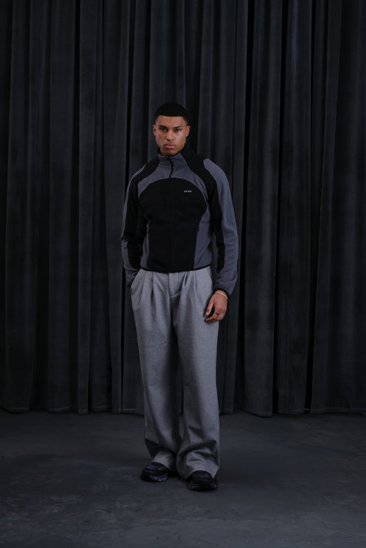 Männer-Sportjacke in Schwarz-Grau mit Color-Block-Design, kombiniert mit grauer Jogginghose vor dunklem Vorhang.