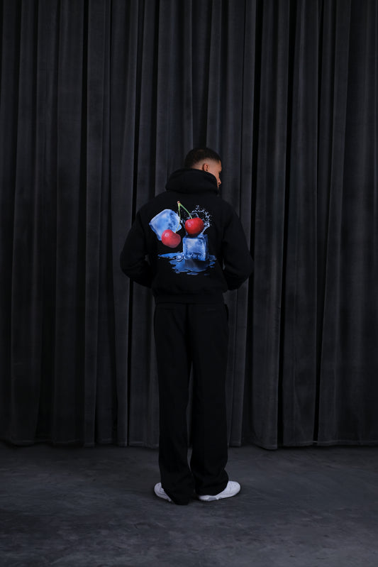 Person in schwarzem Hoodie von hinten mit farbigem Rückenprint (Kirschen, Eiswürfel und Wasser-Splash); schwarze Hose und weiße Sneaker vor dunklem Vorhang.