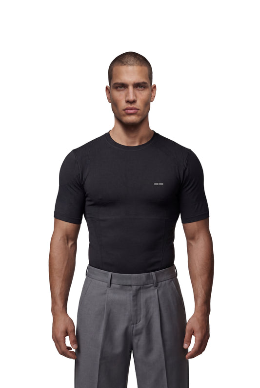 Mann trägt OLAKALA Compression T-Shirt Black, eng anliegend ohne Seitennähte, strukturiertes Design für hohe Bewegungsfreiheit, kombiniert mit grauer, hochtaillierter Hose.