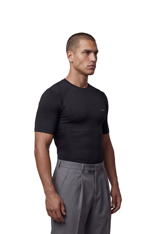 Männliches Model trägt schwarzes Compression T-Shirt von OLAKALA, mit körpernaher Silhouette und nahtloser Konstruktion, kombiniert mit grauen, hoch taillierten Anzughosen.
