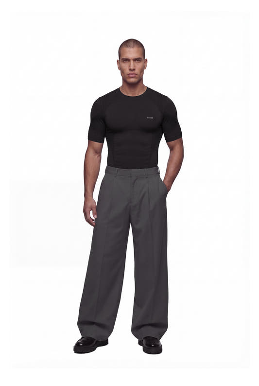 Junger Mann trägt das OLAKALA Compression T-Shirt Black, körperbetont und nahtlos für hohen Komfort, kombiniert mit grauen, weiten Hosen und schwarzen Schuhen.