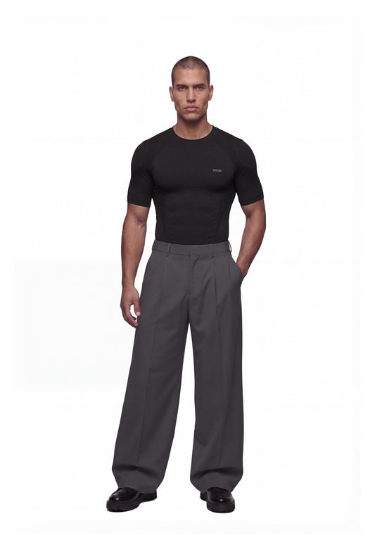 Junger Mann trägt das OLAKALA Compression T-Shirt Black, körperbetont und nahtlos für hohen Komfort, kombiniert mit grauen, weiten Hosen und schwarzen Schuhen.