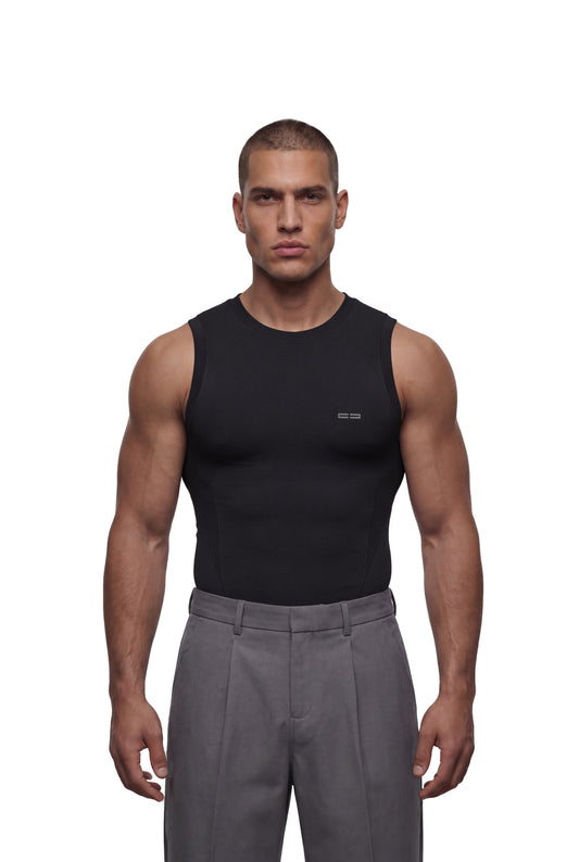 Männermodell trägt OLAKALA Compression Tanktop Black, körpernah, nahtlos, über grauen, hochgeschnittenen Faltenhosen – betont die Performance-Funktionalität und komfortable Passform.