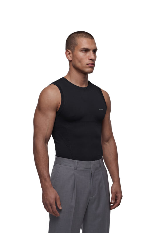 Männermodell im schwarzen OLAKALA Compression Tanktop mit seitlichen Nähten und kleinem Logo, kombiniert mit grauen, hochtaillierten Anzughosen, auf weißem Hintergrund.