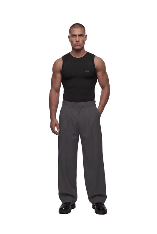Mann trägt OLAKALA Compression Tanktop Black, kombiniert mit grauen, weiten Hosen und schwarzen Lederschuhen, zeigt körpernahe Passform und funktionales Design ohne Seitennähte.