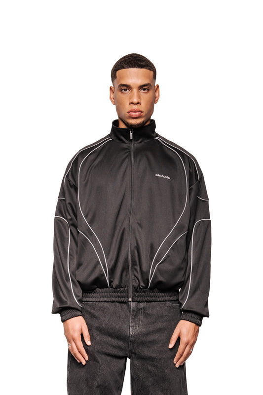 Mann trägt Rave Trackjacket Black mit reflektierenden Paspelierungen, hohem Kragen und gesticktem OLAKALA Logo. Jacke hat elastische Bündchen, Eingriffstaschen, lockerem Schnitt.