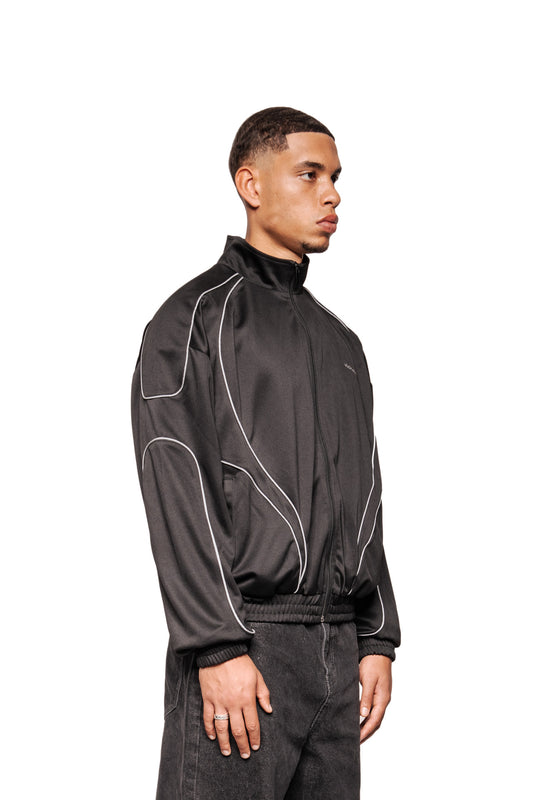 Junger Mann im Profil trägt Rave Trackjacket Black mit Stehkragen, reflektierenden Nähten, elastischen Bündchen und gesticktem OLAKALA Logo.