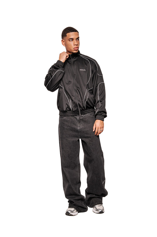 Ein Modell trägt das Rave Trackjacket Black mit Stehkragen, reflektierenden Teilungsnähten und gesticktem OLAKALA Logo, kombiniert mit lässigen Jeans und Sneakers.