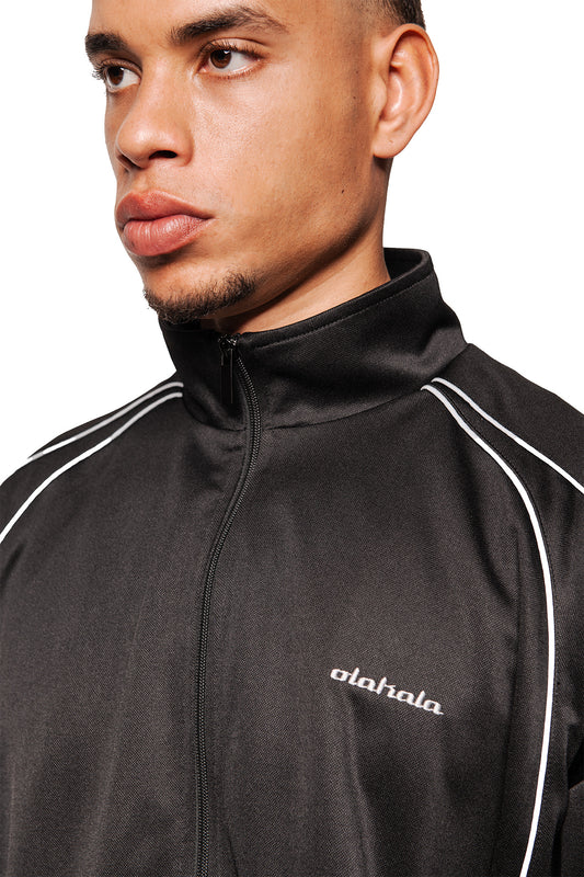 Mann mit schwarzer Rave Trackjacket, Stehkragen, weißer Paspelierung, gesticktem OLAKALA Logo auf Brust, Reißverschluss vorne, unisex Design.
