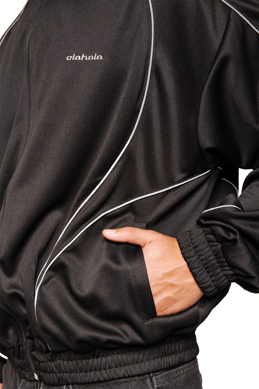 Person trägt Rave Trackjacket Black mit reflektierenden Paspelierungen, gesticktem OLAKALA Logo, Eingriffstaschen und elastischen Bündchen. Hand in Seitentasche, lockere Passform.