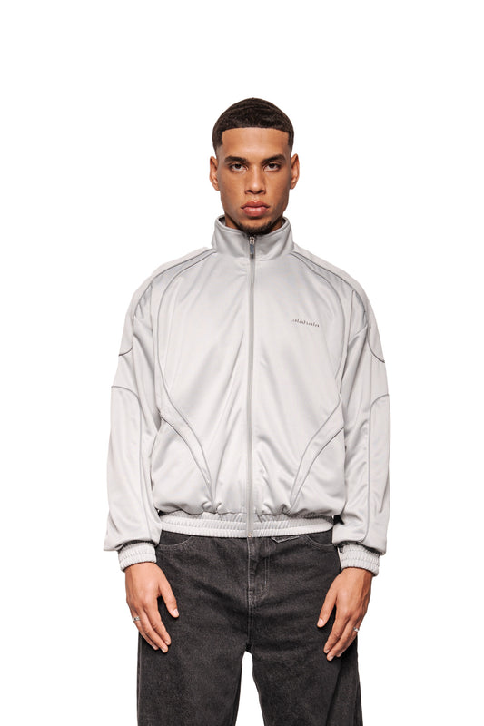Junger Mann trägt den Rave Trackjacket Gray mit Stehkragen, Reißverschluss, reflektierenden Nähten und gesticktem OLAKALA-Logo, kombiniert mit lockeren, schwarzen Jeans.