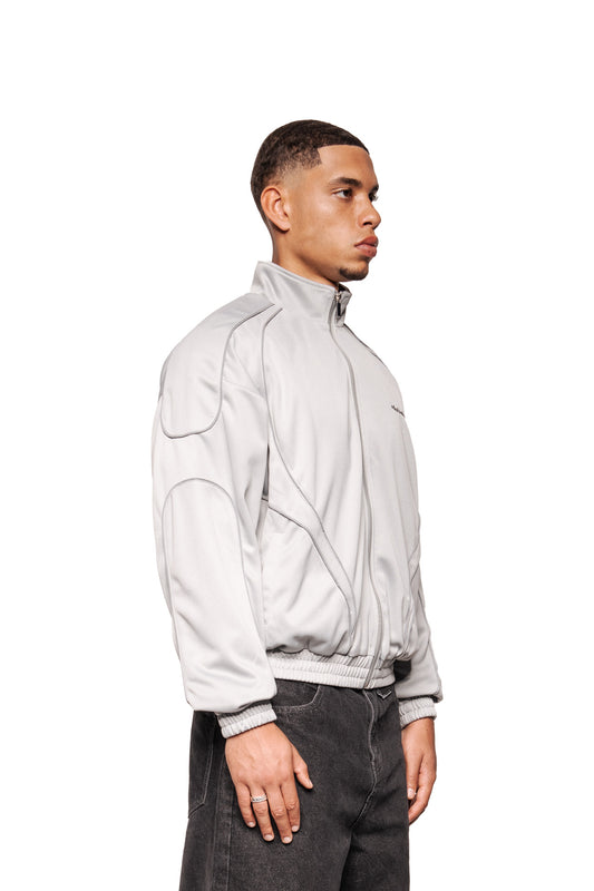 Eine Person trägt die Rave Trackjacket Gray mit Stehkragen, Reißverschluss, reflektierenden Nähten, elastischen Bündchen und gesticktem OLAKALA-Logo auf der Brust.