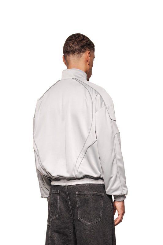 Männliches Modell mit dem Rücken zur Kamera, trägt Rave Trackjacket Gray mit Stehkragen, reflektierenden Teilungsnähten, elastischem Saum und gesticktem OLAKALA-Logo, kombiniert mit weiten Jeans.