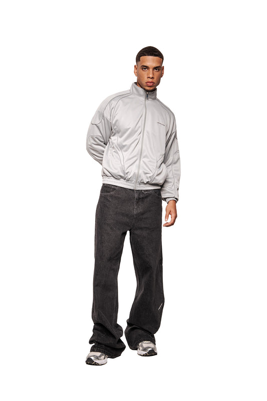 Modell trägt Rave Trackjacket Gray mit hohem Kragen, reflektierenden Nähten, elastischen Bündchen und gesticktem OLAKALA-Logo, kombiniert mit locker sitzenden Jeans und Sneakers.