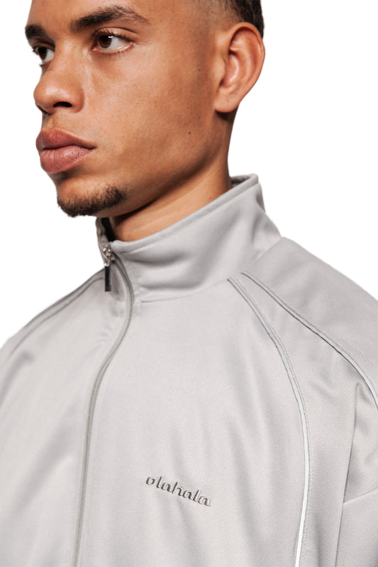 Junger Mann trägt das Rave Trackjacket Gray mit Stehkragen, Reißverschluss, reflektierenden Nähten und gesticktem OLAKALA-Logo auf der linken Brust.